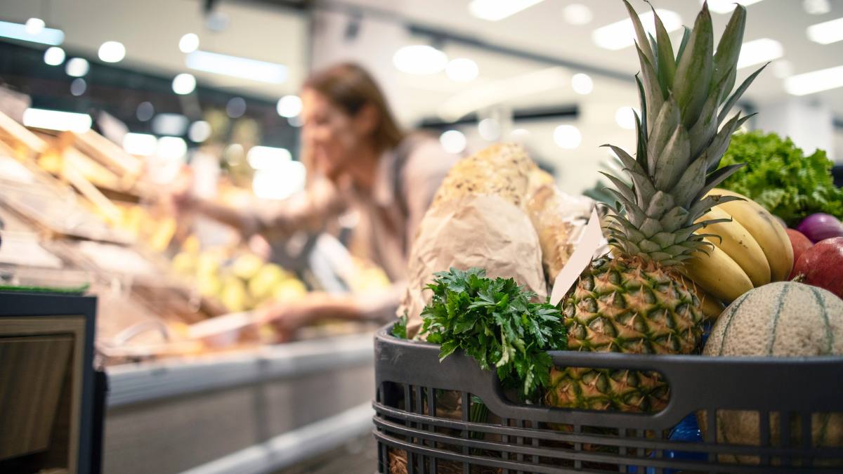 Virus periculos în fructe retragere urgentă. În imagine, un cărucior de supermarket plin cu fructe și în plan depărtat o doamnă la cumpărături