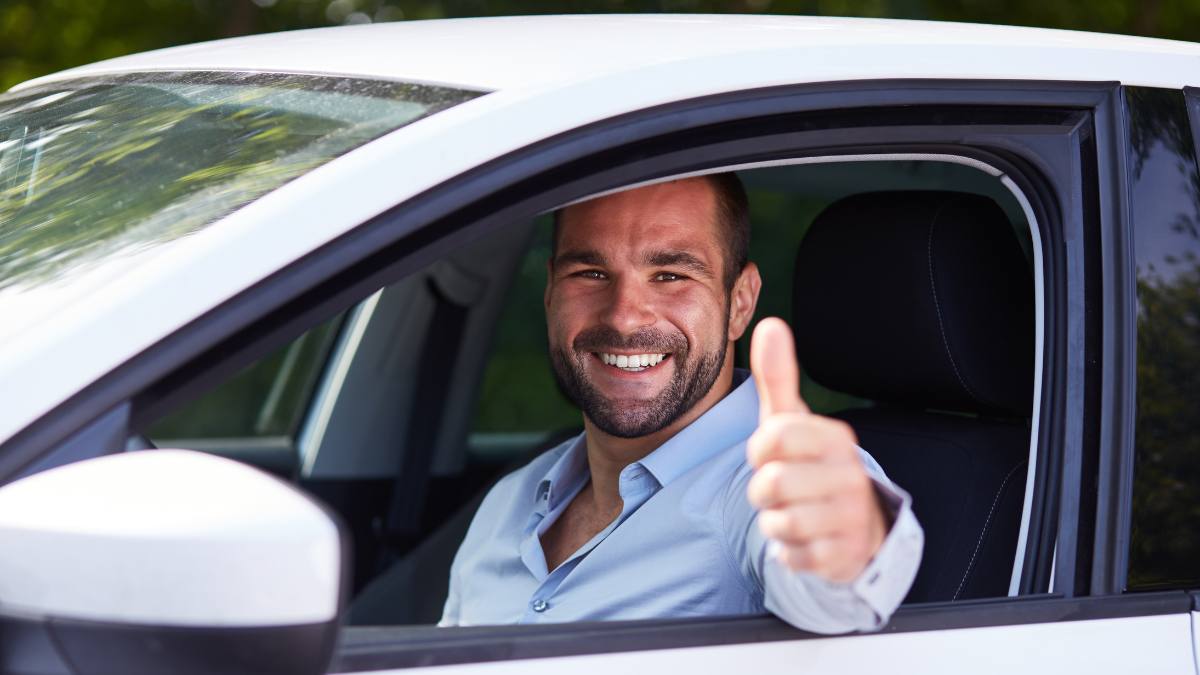 Permis de conducere în România pentru românii din Germania – șofer zâmbitor la volanul unei mașini albe, cu degetul mare ridicat, imagine ilustrativă.