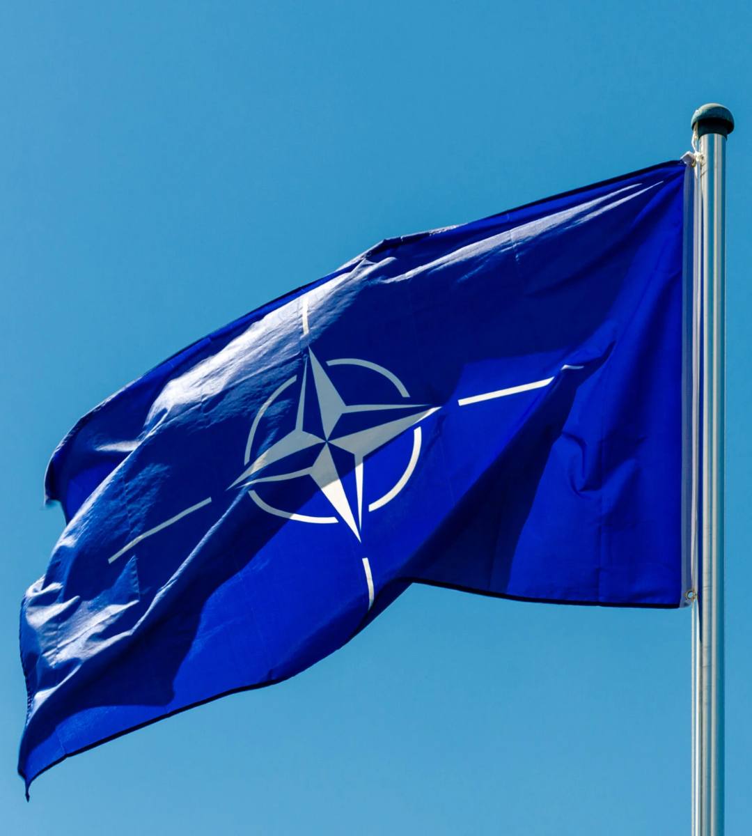 Drapelul albastru al NATO, cu emblema albă a Alianței Nord-Atlantice, flutură pe cer senin, în contextul declarațiilor comandantului suprem despre superioritatea militară față de Rusia.