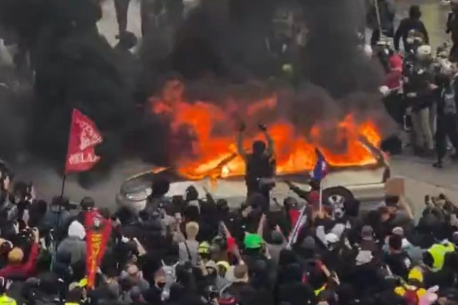 Proteste violente în Europa: o mașină cuprinsă de flăcări, înconjurată de manifestanți, în timpul confruntărilor de stradă.