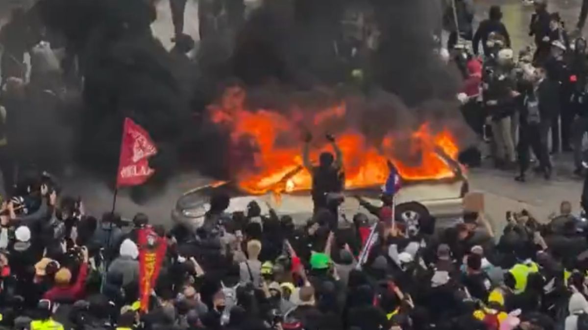 Proteste violente în Europa: o mașină cuprinsă de flăcări, înconjurată de manifestanți, în timpul confruntărilor de stradă.