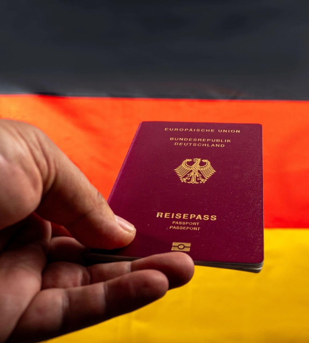 O mână ține un pașaport german în fața drapelului Germaniei, imagine ilustrativă despre cetățenia germană și renunțarea la aceasta.
