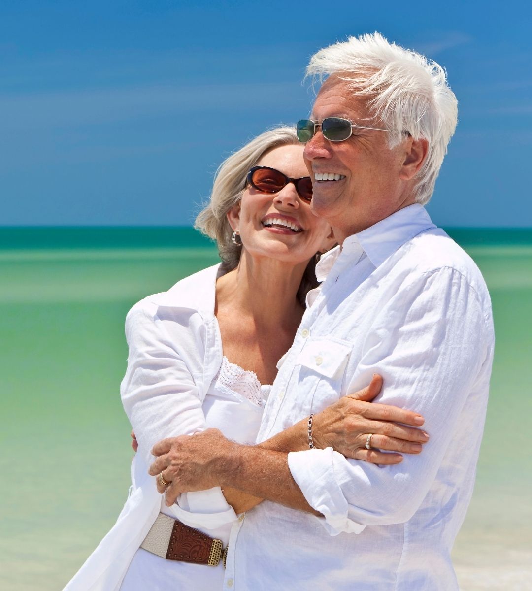 Cuplu de pensionari zâmbitori, îmbrăcați în alb și purtând ochelari de soare, îmbrățișați pe o plajă tropicală, imagine ilustrativă pentru cele mai bune țări în care să trăiești la pensie în 2026.