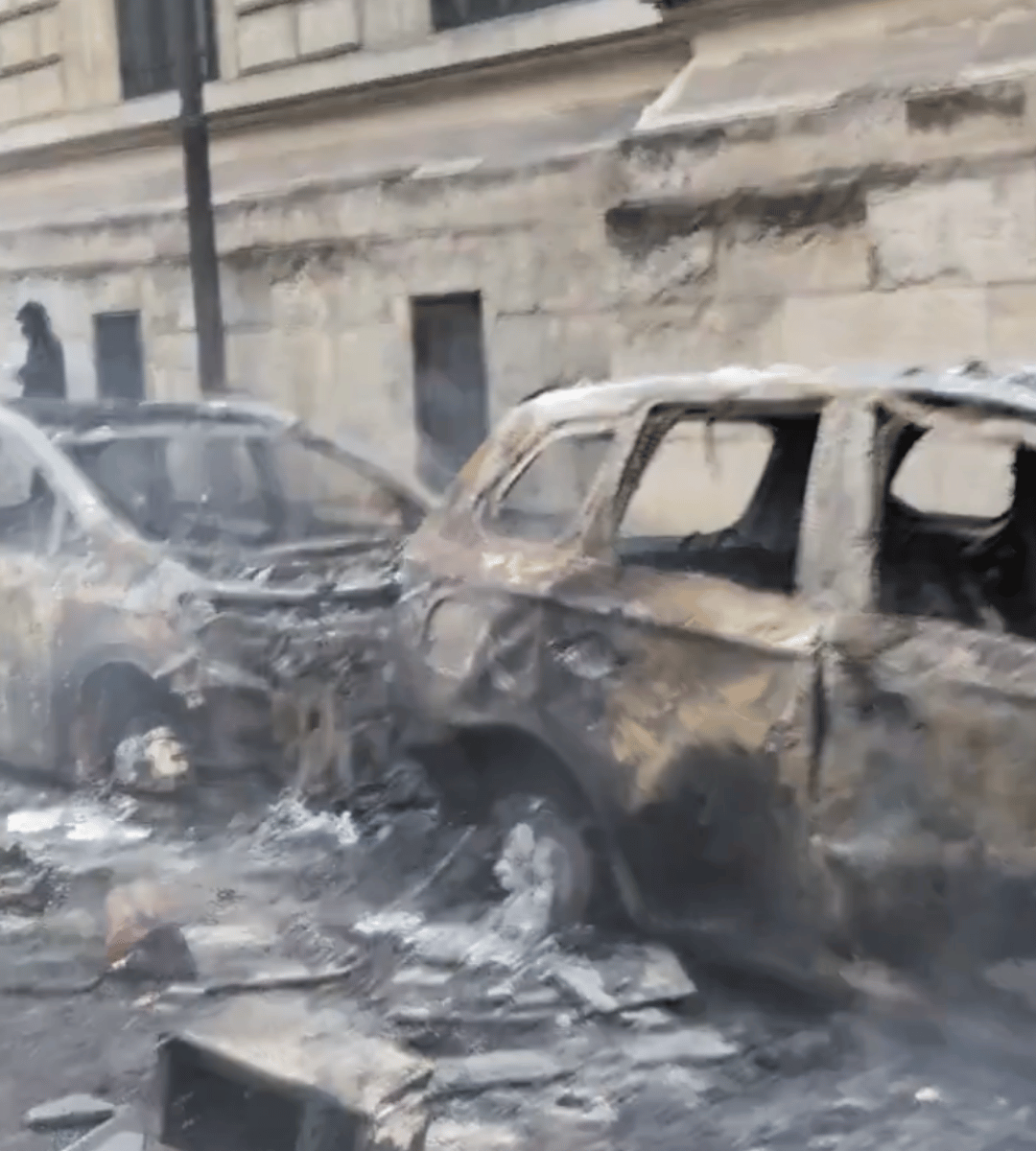 Urmările protestelor violente din Europa: mai multe mașini complet incendiate pe o stradă, printre resturi carbonizate și clădiri avariate.